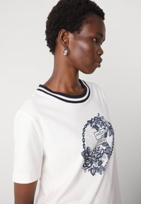 T-shirt bianco in cotone a maniche corte, con colletto a coste blu. Il design frontale include motivi floreali e catene in blu navy.