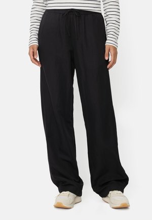 Person trägt lockere schwarze Hose mit Kordelzugbund, weiße Sneakers und weißes Langarmshirt mit schwarzen horizontalen Streifen.