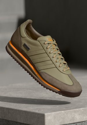 Olijfgroene en bruine sneaker met oranje accenten en Adidas-logo, getoond in de lucht boven een grijze betonnen ondergrond.