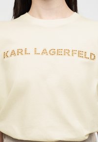 Γυναίκα που φοράει ανοιχτό μπεζ πουκάμισο με κεντημένα γράμματα σε χρώμα ταμπέλα "KARL LAGERFELD" στο ύψος του στήθους.