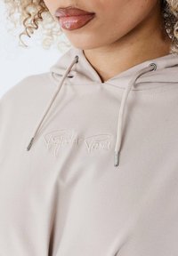 Beige hoodie med snoretræk med sølvspidser, broderet "Project Paris" logo på forsiden, glat tekstur og afslappet design.