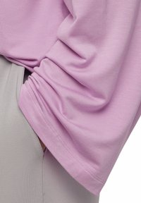 Helles lila Langarmshirt aus weichem, strukturiertem Baumwollstoff mit lässiger Passform. Grauer Stoff ist teilweise darunter sichtbar.