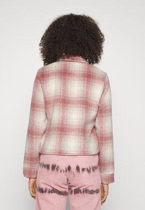 Osoba z kręconymi włosami, ubrana w różowo-białą kraciastą kurtkę i różowe dżinsy z czarnymi wzorami tie-dye, widziana od tyłu.