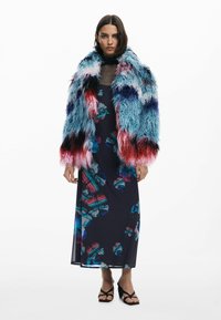 Bunt gefärbter Kunstfellmantel mit langen Ärmeln und lebhaften blauen, rosa und roten Texturen. Darunter ein marineblaues Kleid mit floralen Mustern.