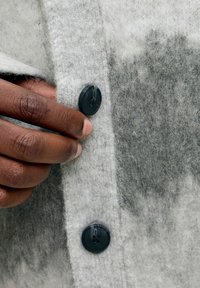 Šedý vlněný kabát s texturovaným povrchem, který má černý knoflíkový detail a částečně rozepnuté zapínání, a odhalenou ruku, která drží látku.