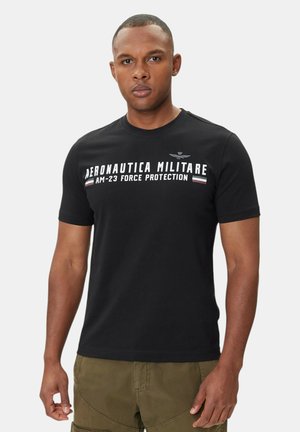 Uomo che indossa una maglietta nera Aeronautica Militare e pantaloni verde oliva, in piedi davanti a uno sfondo semplice grigio chiaro.