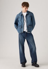 Jonge man in een blauwe denimjacket en bijpassende wijde jeans, wit T-shirt en zwarte schoenen, staande tegen een effen achtergrond.