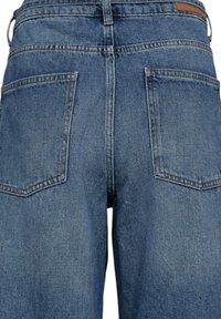 Jean en denim à teinte moyenne avec une coupe décontractée, doté de deux poches arrière et d'une ceinture propre avec un accent en cuir marron clair.