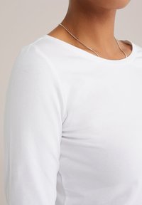 Oberkörper einer Person, die ein schlichtes weißes Langarmshirt und eine dünne silberne Kettenhalskette vor einem neutralen Hintergrund trägt.