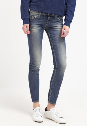 Jeans ajustados azules de mezclilla, con un lavado deslavado, bolsillos estándar y combinados con zapatillas blancas; textura suave y ajuste ceñido.