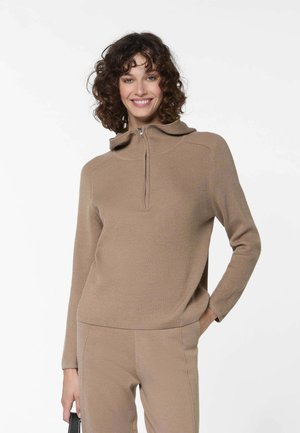 Lachende vrouw met krullend haar, gekleed in een beige geribbelde sweater met rits en bijpassende beige broek, met in één hand een zwarte tas.