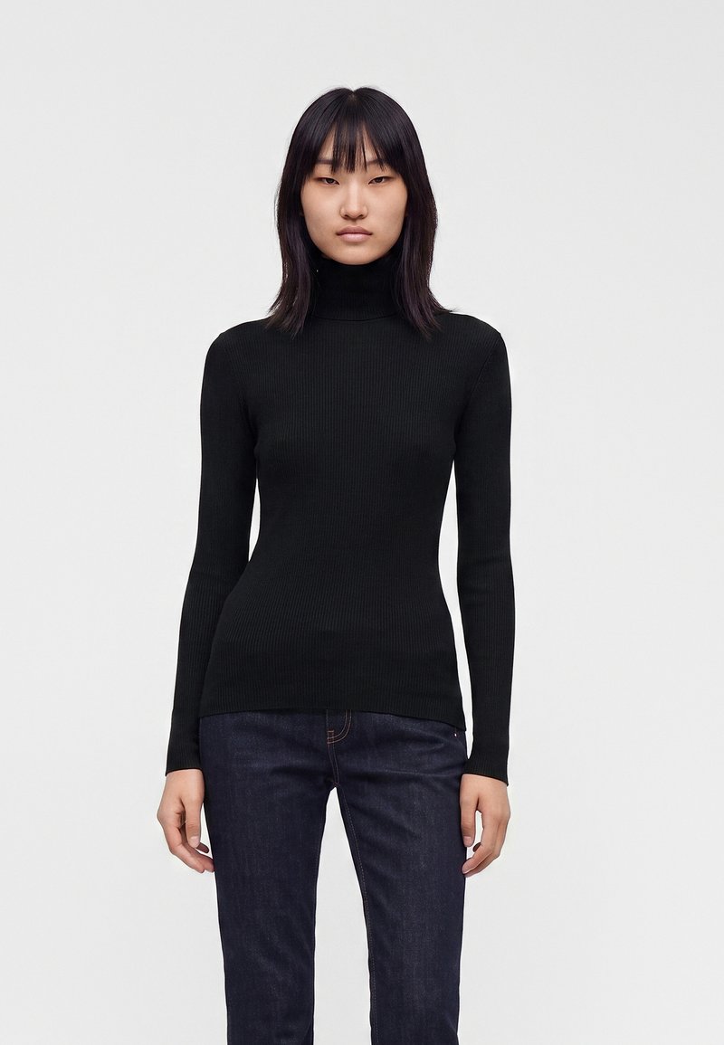 Femme portant un pull à col roulé côtelé noir ajusté et un jean bleu foncé, debout devant un fond clair uni.