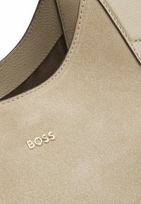 Borsa a tracolla in camoscio beige con un accento in pelle liscia, con dettaglio logo dorato. La texture è morbida e include cuciture sottili.