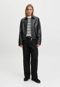 Persona con cabello rubio hasta los hombros, vistiendo una chaqueta de cuero negra, pantalones negros, un suéter a rayas en blanco y negro, y zapatos negros, contra un fondo blanco.