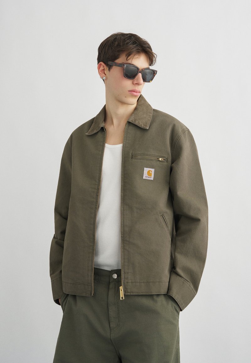 Giovane uomo con occhiali da sole scuri e giacca verde oliva sopra una maglietta bianca, in piedi davanti a uno sfondo chiaro e semplice.