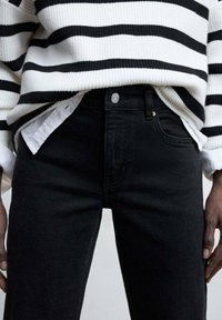 Vaqueros de mezclilla negros con un ajuste de cintura alta, presentan un cierre de botón delantero, bolsillos laterales y sutiles detalles de costura, combinados con un suéter a rayas.