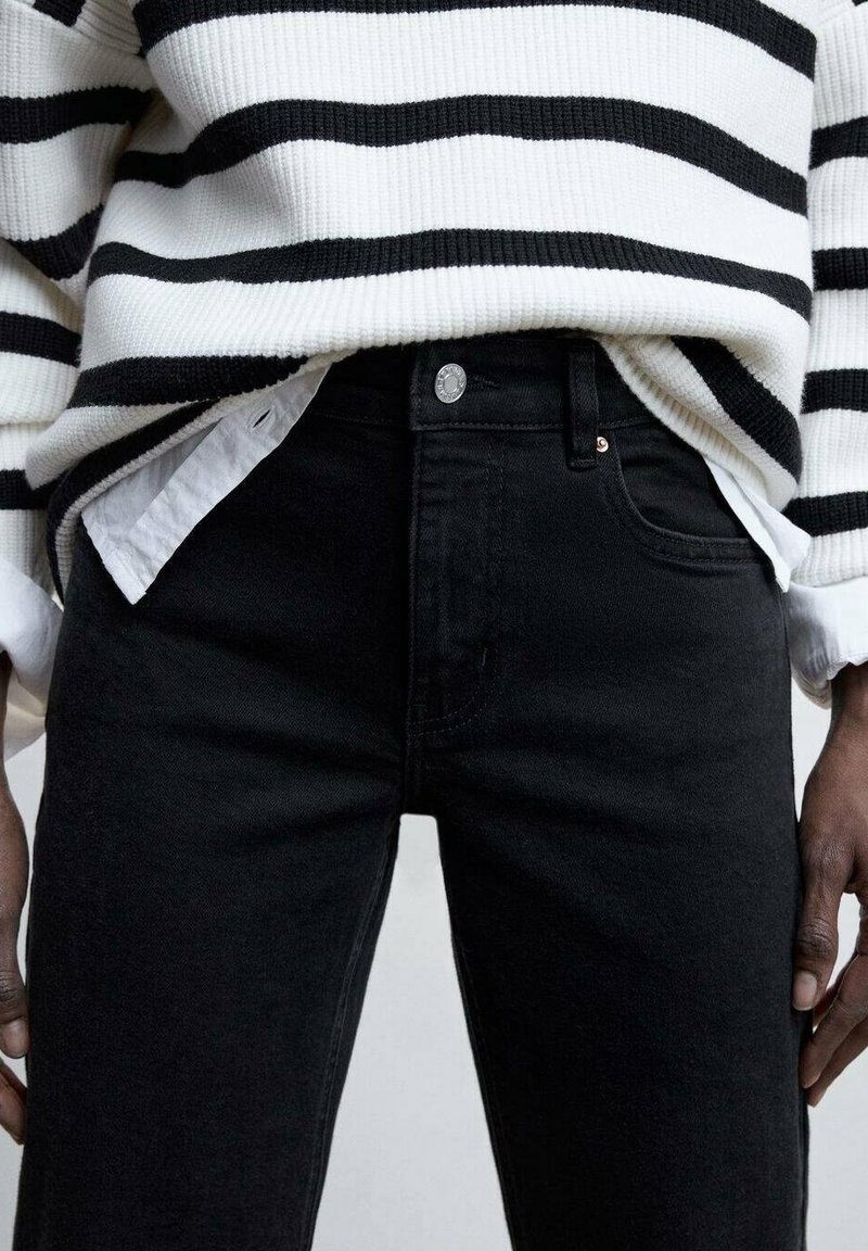 Vaqueros de mezclilla negros con un ajuste de cintura alta, presentan un cierre de botón delantero, bolsillos laterales y sutiles detalles de costura, combinados con un suéter a rayas.