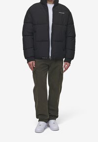 Sort dunjakke med høj krave, lynlåslukning og ribstrikkede ærmeender, kombineret med olivengrønne cargobukser og hvide sneakers.