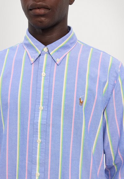 Chemise bleu à boutons avec des rayures verticales roses et vertes, ornée d'un petit logo de polo brodé marron sur la poitrine. Tissu en coton.
