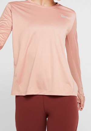 Kobieta w jasnoróżowej sportowej bluzce z długim rękawem i otworami na kciuki oraz ciemnoczerwonych legginsach na jednolitym tle.