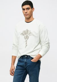 Felpa bianca con un design astratto e testurizzato in beige e grigio. Dotata di maniche lunghe e colletto rotondo, abbinata a jeans blu.