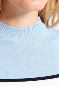 Detailní záběr na osobu, která má na sobě světle modrý pletený svetr s tmavým horizontálním pruhem blízko spodního okraje.