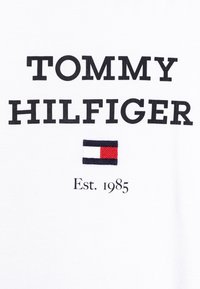 Bílý tričko s výrazným černým textem "TOMMY HILFIGER," malou vyšívanou nášivkou s červenými, bílými a modrými pruhy a nápisem "Est. 1985" pod ním.