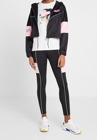 Zwarte cropped jack met roze mouwen, grafisch wit t-shirt en goed aansluitende zwarte leggings met witte zijkanten. Paar witte sneakers.