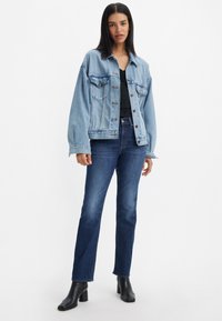 Giacca in denim azzurro chiaro con due tasche sul petto e jeans blu scuro. Modello che indossa stivaletti neri con tacco, in piedi su uno sfondo neutro.