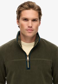 Zielony polarowy sweter z wysokim kołnierzem, złotym zamkiem błyskawicznym i granatowymi wykończeniami. Charakteryzuje się jednolitą strukturą i minimalistycznym designem.