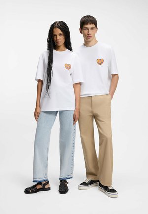 Donna e uomo in piedi fianco a fianco, entrambi con T-shirt bianche con logo a forma di cuore sorridente arancione; donna in jeans chiari e sandali, uomo in pantaloni beige e scarpe da ginnastica.