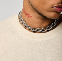 Choker en métal argenté avec des maillons entrelacés, porté sur un vêtement texturé beige clair, accentué par des tatouages sur le cou.