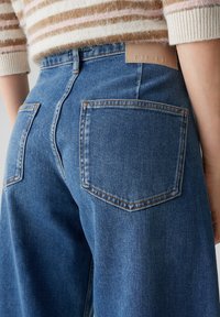 Jean en denim bleu clair à coupe décontractée, avec deux poches arrière, des coutures contrastantes et un patch en cuir portant la mention "OPUS" à la ceinture.