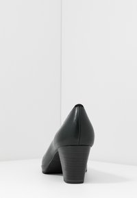 Black leather mid-heel shoe with stacked heel, shown from the back on a white surface against a white corner background.