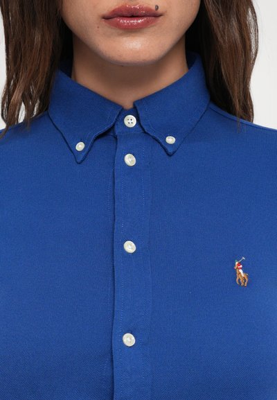 Zils polo krekls ar apkakli un baltiem pogām, kurā ir neliels izšūts zirga un jātnieka logos kreisajā krūtī. Tekstūrēta auduma.