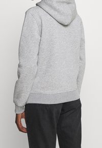 Persona che indossa un felpa con cappuccio grigia chiara e jeans neri, girata di spalle rispetto alla macchina fotografica su uno sfondo chiaro e semplice.