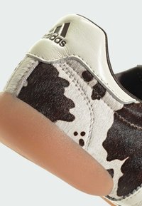 Adidas sneaker featuring a cowhide patterned upper in black and white, with a smooth white leather heel and beige rubber sole.