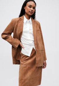 Vrouw in een bruine leren blazer en rok met een wit overhemd, staat met een hand op haar heup tegen een effen lichte achtergrond.
