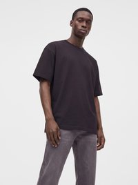 Homme portant un t-shirt noir uni oversize et un jean gris délavé, debout devant un fond gris clair.