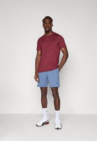 Bordeauxrood shirt met korte mouwen en een subtiel textuurpatroon, gecombineerd met lichtblauwe shorts. Het model draagt witte sneakers en sokken.