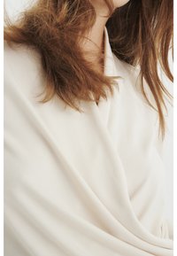 Kvinnas midja med vågigt brunt hår som bär en krämfärgad, omslagstyp blus eller klänning mot en vit bakgrund.