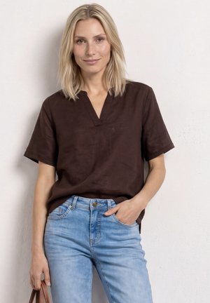 Blondynka ubrana w ciemnobrązową bluzkę z krótkim rękawem i jasnoniebieskie dżinsy, stojąca na tle jednolitej jasnej ściany, delikatnie uśmiechająca się.