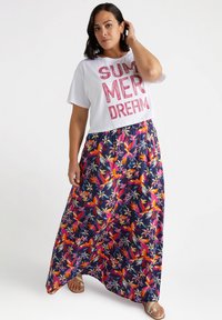 T-shirt bianco tagliato con testo "SUMMER DREAM", abbinato a una lunga gonna blu marina con vivaci motivi floreali e uccelli. Sandali con punta aperta.