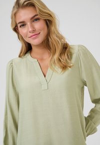 Light green, textured blouse with a v-neckline, long sleeves, and subtle gathered detailing at the shoulders. Soft, flowing fabric.