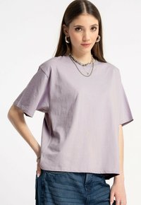 Młoda kobieta w oversize'owej jasnofioletowej koszulce, warstwowych srebrnych naszyjnikach, kolczykach w kształcie kół i niebieskich dżinsach, stoi z ręką na biodrze.