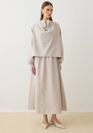 Donna che indossa un lungo abito beige chiaro con maniche a sbuffo e un sovrapposto smanicato ampio, abbinato a scarpe crema a punta con tacco basso.