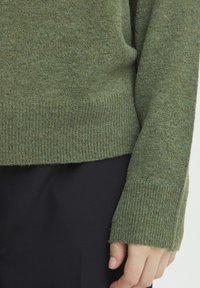 Grüner gestrickter Pullover mit gerippten Bündchen und Saum, der eine weiche Textur aufweist. Der Ärmel ist etwas länger und hat eine lockere Passform. Über schwarzem Stoff getragen.