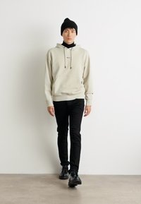 Sudadera beige clara con un bolsillo frontal y cordones negros, combinada con pantalones ajustados negros y zapatos negros, llevada bajo un gorro negro.