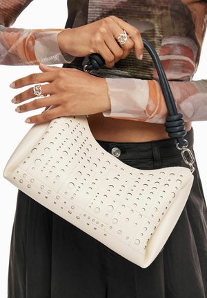 Ecrufarbenes Lederhandtasche mit durchbrochenen Mustern, geschwungener Form, schwarzem Griff mit Knoten-Detail, metallischen Beschlägen und Reißverschluss.
