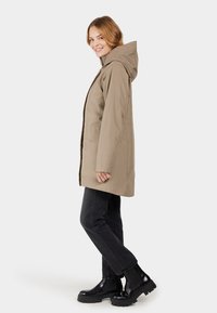 Beige wasserdichte Jacke mit Kapuze, im geraden Schnitt, Seiten-taschen, kombiniert mit schwarzen Jeans und klobigen schwarzen Stiefeln.
